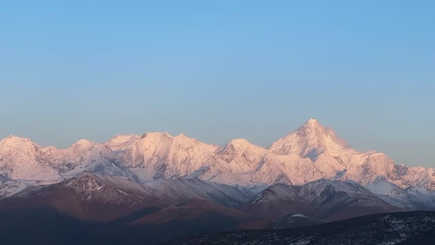The majestic snow-capped peaks of Gongga Mountain (Minya Konka) glow with warm light during sunrise in Sichuan, China. This stunning natural landscape features dramatic mountain ranges and a clear