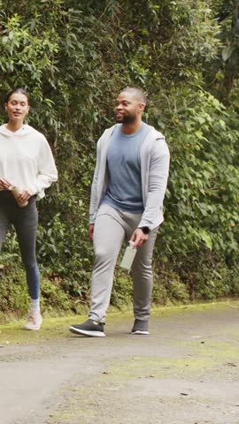 Vertical video: Walking sporty duo pausing on seeing plant on moss path, checking leaf with bottle. Nature, trail, greenery, outdoors, pair, exploration, vertical video
