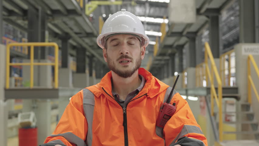 Man in orange high-visibility jacket and white hard hat uses radio at Train Depot. Industrial machinery surrounds him within dark warehouse environment, conveying operational oversight and diligent monitoring of equipment maintenance. Focused attention suggests preparedness in this technologically advanced depot setting.