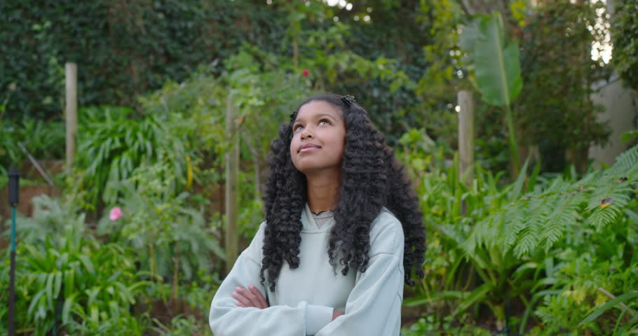African American teenage female standing arms crossed in hoodie in yard reacting to camera smiling. Garden, outdoors, greenery, plants, nature, candid, happiness