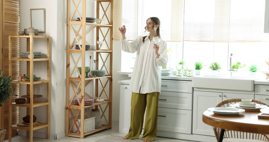 Happy young woman putting clean glasses onto shelving unit in kitchen