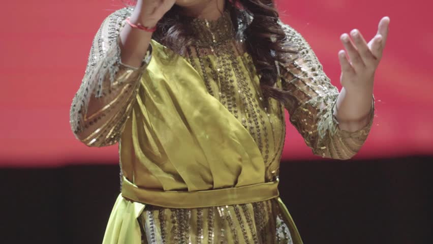 Close Up Video of an Unrecognizable Asian Young Female Singer Gesturing with Her Hands on Stage During a Live Concert. Wearing a Glamorous Gold Sequined Dress with a Draped Satin Sash. Highlighting Fabric Detail, Textile Texture, and Authentic Fashion.