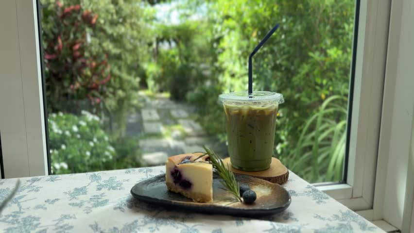 A refreshing iced matcha latte and delicious blueberry cheesecake served on a cozy table by a window, with a serene garden view.