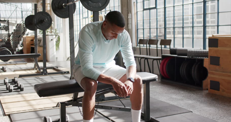 African American man pausing between sets on flat bench at gym, checking watch, gazing at barbell. Athlete, strength, weights, equipment, warehouse, sunlit, industrial