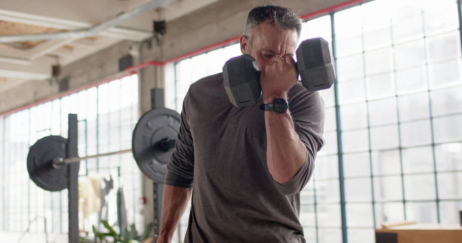 Middle-aged man holding hex dumbbells, curling, lowering to bench at gym, preparing for next set. Athlete, training, strength, weights, barbell, studio, focus