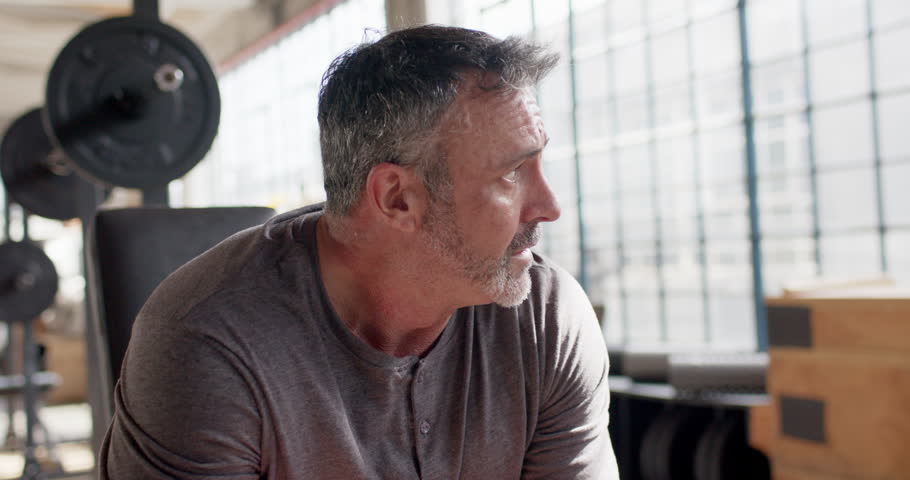Middle-aged man sitting on bench by barbell in gym loft, hearing off-camera cue, turning to camera. Lifting, focus, gaze, industrial, daylight, studio, fitness
