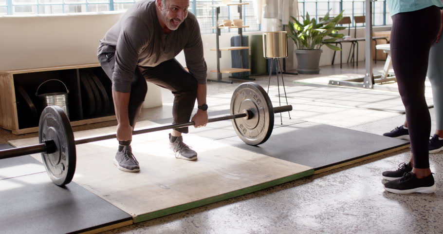 Trainer cueing, male lifter bending, gripping barbell on platform, pressing up for strength in loft. Gym, weightlifting, coaching, studio, daylight, industrial, focus
