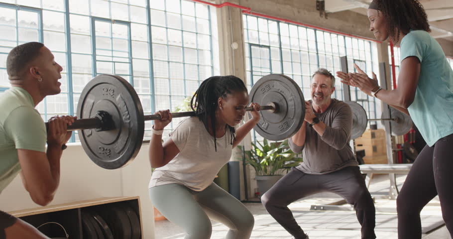 Diverse group, coach clapping, African American woman squatting barbell pressing overhead in gym. Lifting, weights, studio, industrial, sunlit, teamwork, spotting
