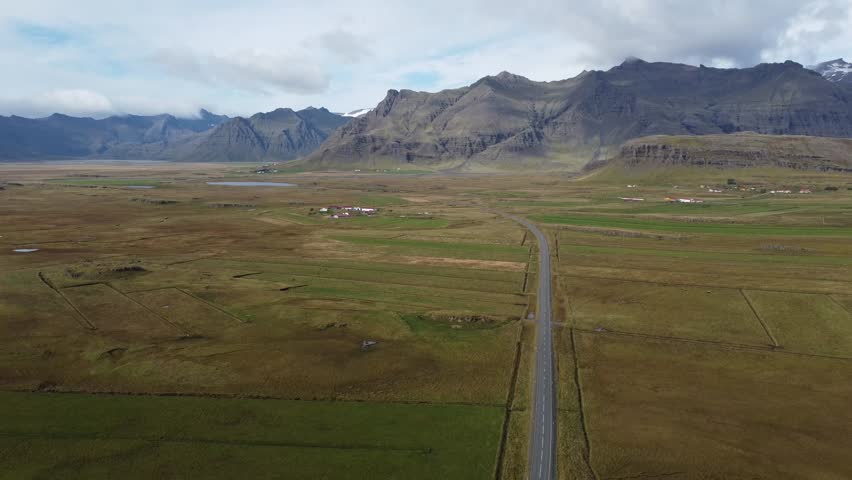 Aerial View of Straight Road Through Icelandic Valley and Mountains - Drone view of a straight asphalt road cutting through vast fields toward rugged mountains under overcast skies.