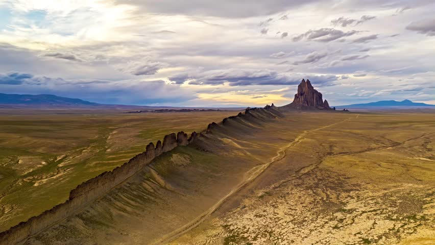 Cinematic drone footage of iconic Shiprock rock formation in New Mexico. Vast desert plains under dramatic cloudy sky. Natural wonder, geological formation.