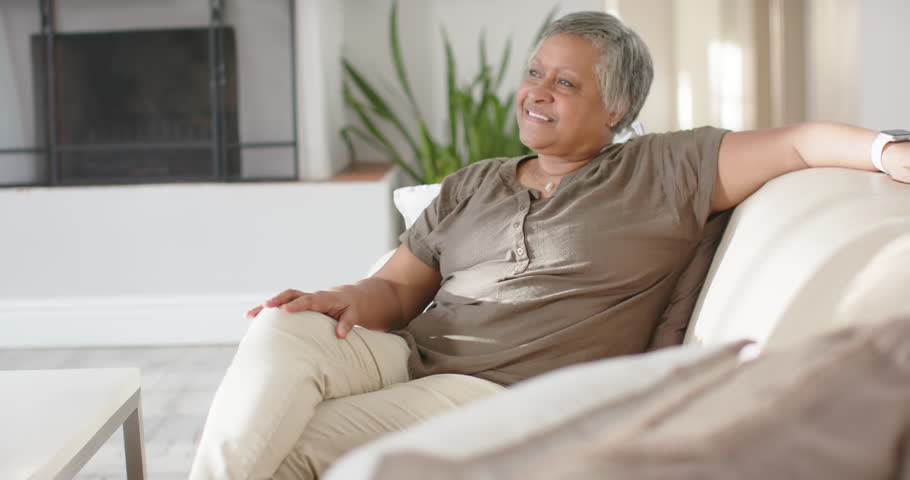 Senior African American female responding to off-camera voice, turning and smiling on sofa at home. Older, woman, interior, room, plant, fireplace, natural