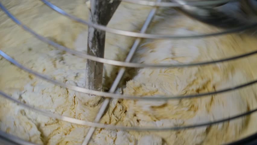 Dough is being kneaded inside an industrial mixer bowl. Close-up view showing the mixing, kneading and preparation process for bread, flatbread, pizza and pastry production.