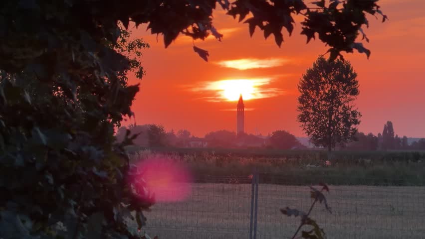 Dramatic red sunset over a rural landscape. With the sun setting behind a distant church steeple. The warm evening light illuminates the countryside fields. Creating a peaceful and serene atmosphere
