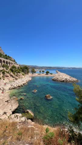 Pebble beach at Plage de l’Estaque in Marseille with vivid blue Mediterranean waters and the iconic railway viaduct above. Scenic rocky coastline in bright sunlight.
