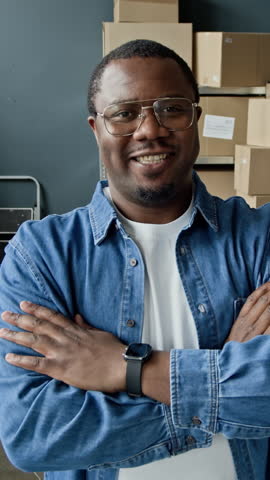 Portrait of African American young adult male warehouse worker standing with arms crossed and smiling. Footage is ideal for e-commerce and logistics marketing, vertical