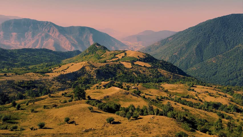 A serene landscape of rolling hills and mountains covered with trees and grass, set against a backdrop of hazy mountains under a pinkish sky at sunset