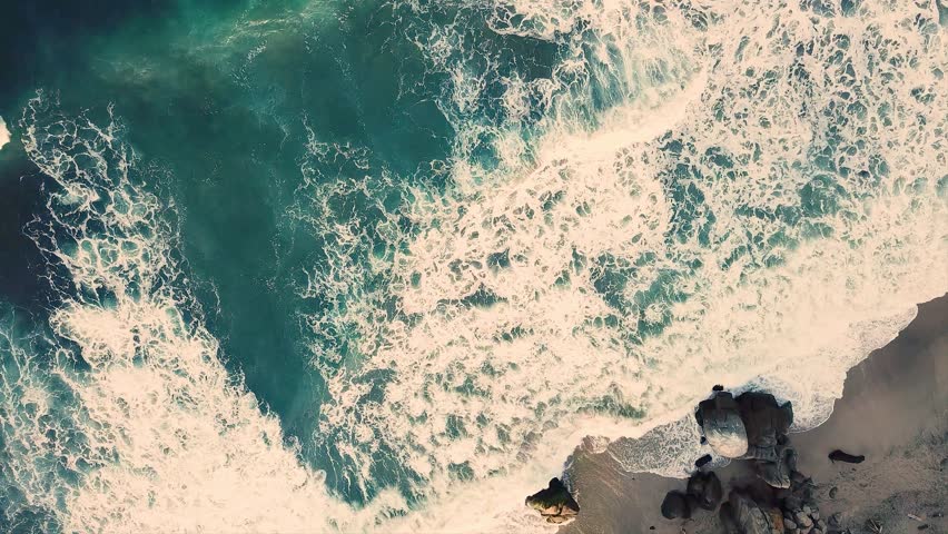 Aerial drone shot of turquoise ocean waves with white foam crashing on rocky shoreline and sandy beach with large boulders