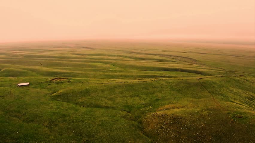 Aerial view of rolling green hills with a small barn and fence at sunset, casting a warm orange glow over the scene