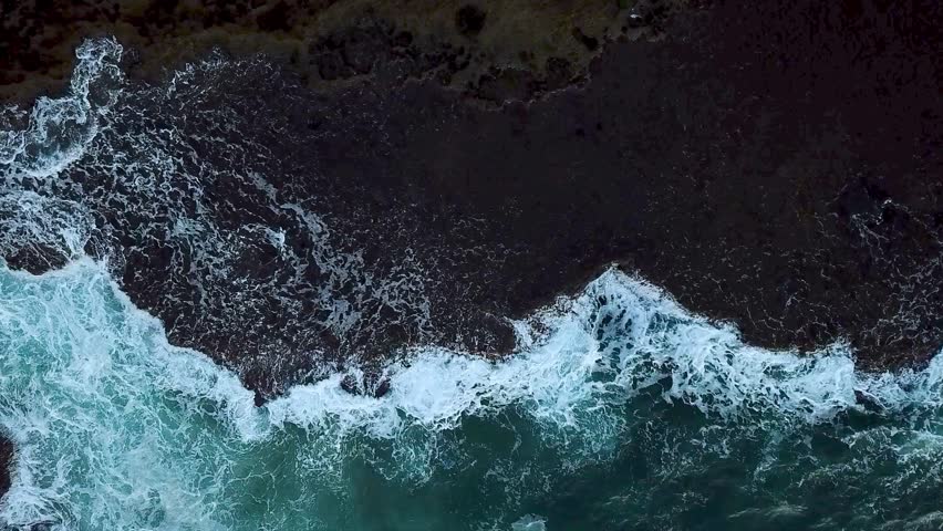 Aerial drone view of large ocean waves crashing on a dark rocky shoreline with white foam and turquoise water