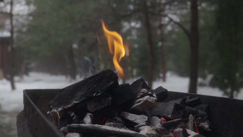 Extreme close-up shot of bright orange flame burning on charcoal inside a metal fire pit. Small fire and light smoke rising in winter forest environment. Shot in slow motion with shallow depth of field.