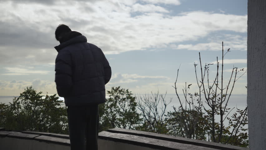 Man sitting on a viewpoint terrace with a panoramic sea view on a cloudy day. The person is facing the camera but looking away, creating a calm and contemplative mood. Natural outdoor scene with sea, sky and coastal vegetation in the background.