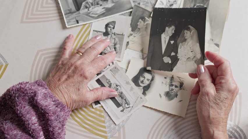 Wrinkled hands of a senior woman touching old black and white photographs of her youth, reminiscing about past memories, her wedding day, and her husband who was a soldier in the army