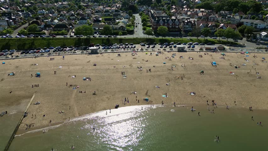 People are on Colwyn Bay Beach in North Wales. Some walk along the shore while others sunbathe or swim in the water. Cars are parked nearby and houses are visible in the background.