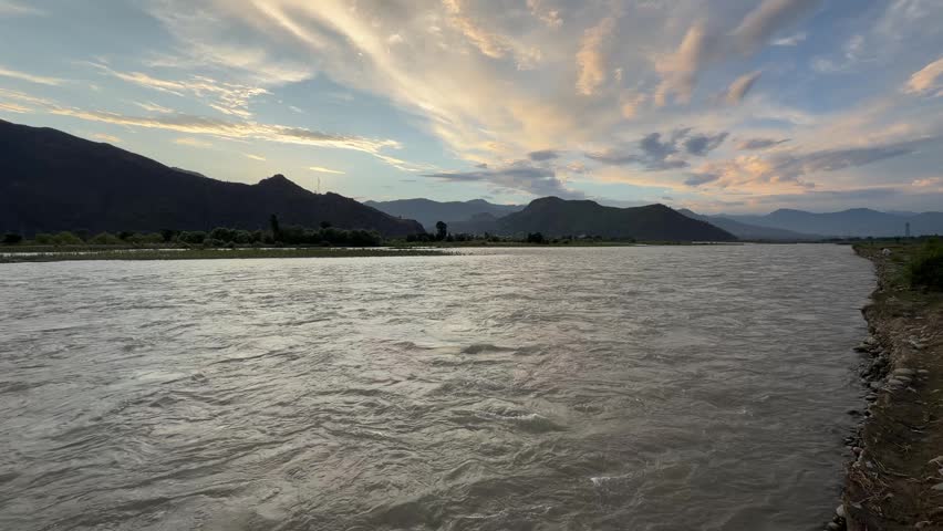 A wide, fast-flowing river cuts through a mountainous valley beneath a expansive, cloud-streaked sunset sky.