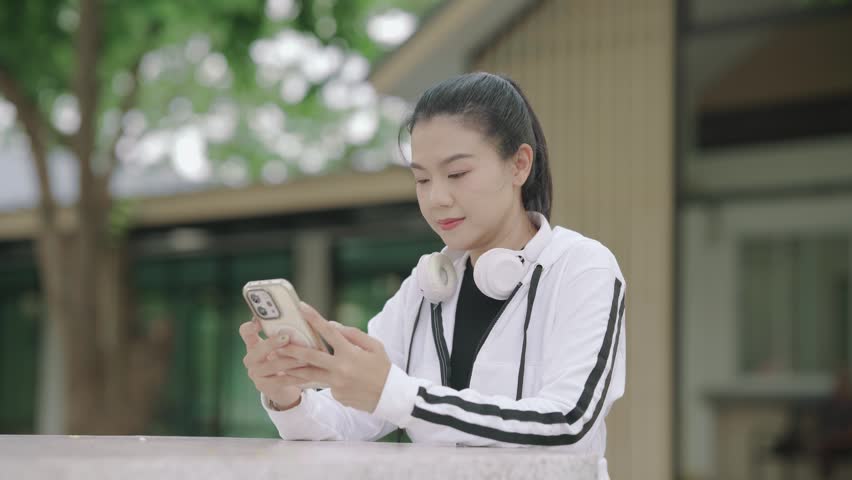 Young Asian woman using a smartphone outdoors after exercise while wearing wireless headphones around her neck. Mobile technology, communication, healthy lifestyle, fitness, and digital lifestyle concept.
