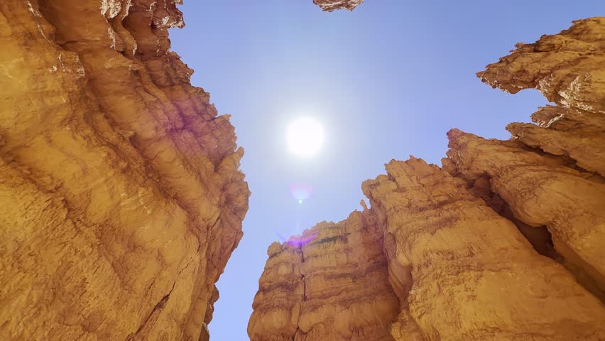 POV Tilt Up to Red Rock Spires and Bright Sun in Blue Sky in Bryce Canyon Utah. A smooth low angle cinematic shot looking up at surreal red sandstone peaks in Bryce Canyon, Utah. Majestic rock shapes under a bright sun and blue sky create an alien planet atmosphere. USA.