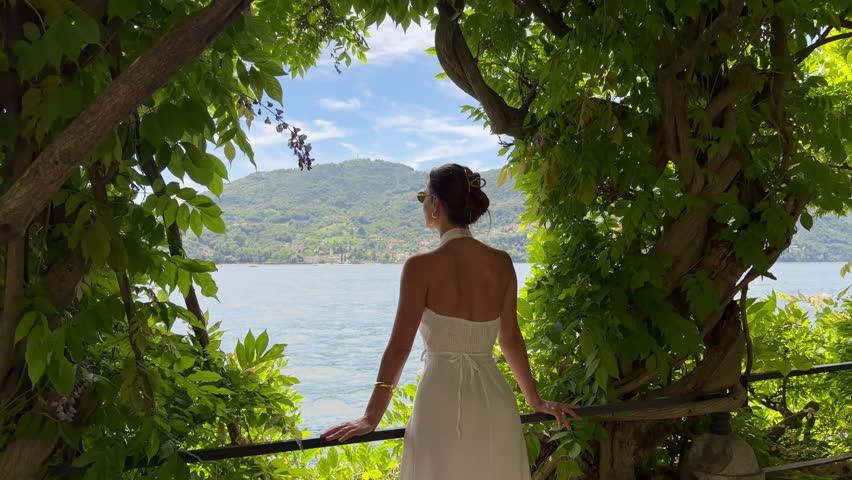 Young beautiful woman in a white dress stands by Lake Como, Italy, as the camera pans to reveal a panoramic view of the lake and mountains during a luxury summer vacation. A travel and lifestyle shot transitioning into a scenic nature landscape in Lombardy, Italy.
