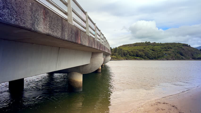 A wide, scenic panoramic view of the modern Pont Briwet concrete bridge running alongside the tidal estuary of the River Afon Dwyryd with lush green hills and rolling mountains, Gwynedd, North Wales, UK