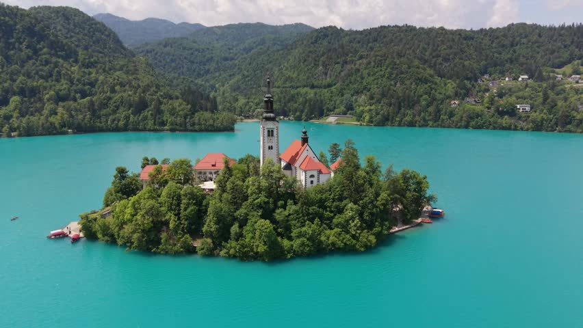 A church stands on an island in Lake Bled. The water is blue and clear and mountains are in the background.