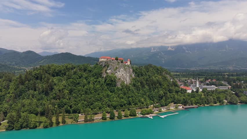 A view of Bled with its castle situated on a hill overlooking the lake and surrounding mountains. People enjoy the scenery by the water.