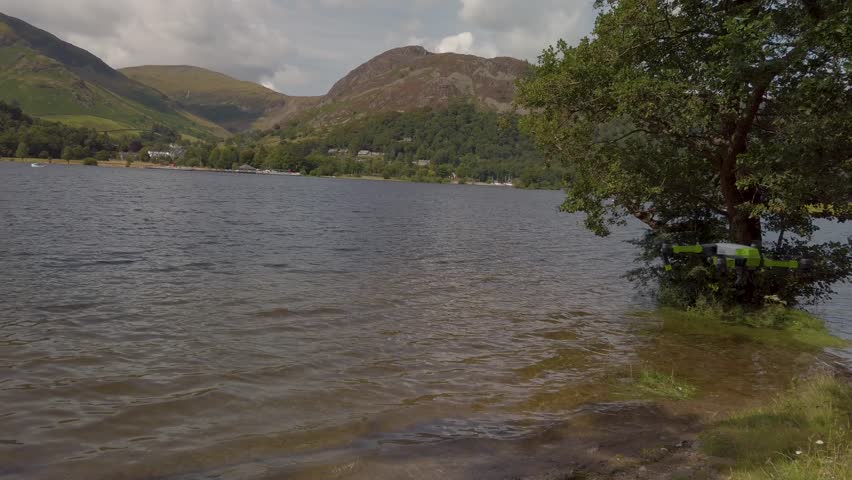 Drone captured footage flying over serene lake landscape. Hills and trees frame scenic view. Drone is visible in flight.