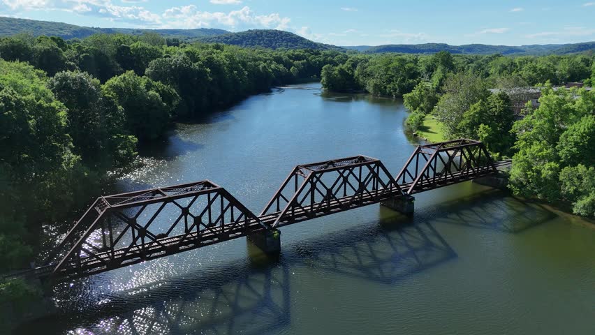 Scenic high-angle aerial drone footage capturing the historic charm of New Milford, Connecticut, located in Litchfield County. The video showcases the beautiful New England suburban landscape, featuring the winding Housatonic River, lush green hillsides, and the classic downtown village green area with its traditional architecture and local streets. Perfect for regional tourism marketing, commercial real estate videos, New England travel documentaries, autumn or summer seasonal concepts, and small-town American lifestyle stock media. Railroad bridge.