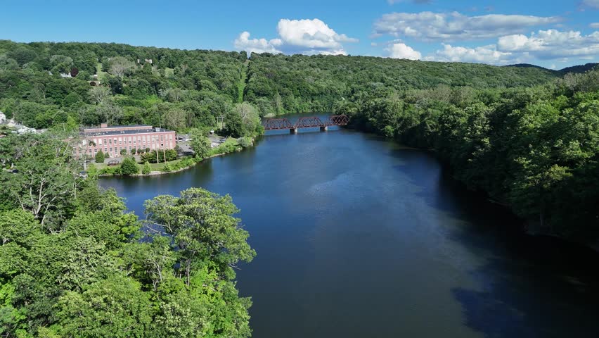 Scenic high-angle aerial drone footage capturing the historic charm of New Milford, Connecticut, located in Litchfield County. The video showcases the beautiful New England suburban landscape, featuring the winding Housatonic River, lush green hillsides, and the classic downtown village green area with its traditional architecture and local streets. Perfect for regional tourism marketing, commercial real estate videos, New England travel documentaries, autumn or summer seasonal concepts, and small-town American lifestyle stock media. Railroad bridge.