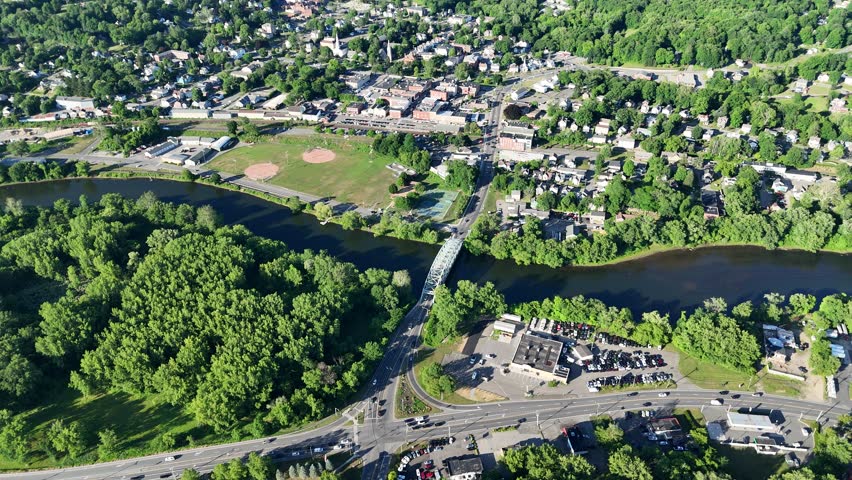 Scenic high-angle aerial drone footage capturing the historic charm of New Milford, Connecticut, located in Litchfield County. The video showcases the beautiful New England suburban landscape, featuring the winding Housatonic River, lush green hillsides, and the classic downtown village green area with its traditional architecture and local streets. Perfect for regional tourism marketing, commercial real estate videos, New England travel documentaries, autumn or summer seasonal concepts, and small-town American lifestyle stock media.