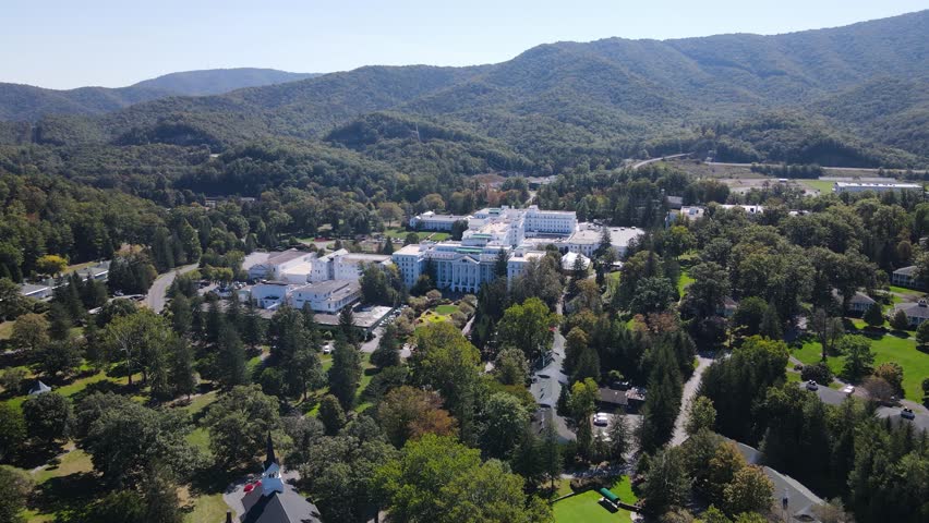Cinematic aerial drone footage capturing historic white resort architecture surrounded by lush green forests and rolling mountains under bright blue sky on sunny autumn day in Virginia.