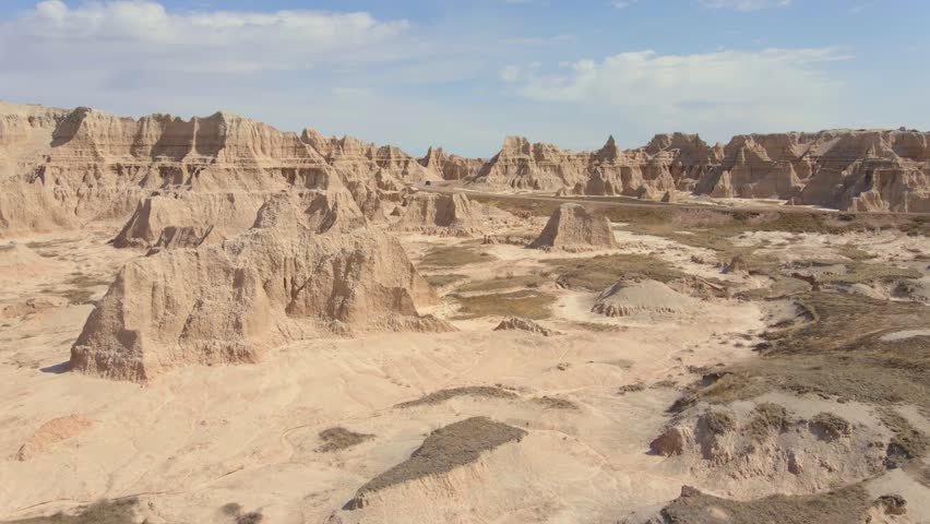 Cinematic aerial footage captures white car traveling along curved asphalt road through rugged rocky terrain and deep canyons within Badlands National Park wilderness landscapes.