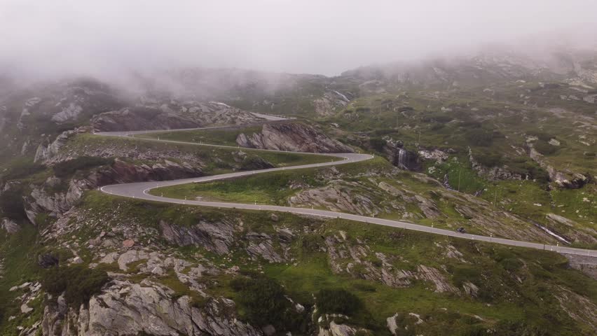 Aerial drone cinematography capturing sweeping mountain landscape with zig zag road in Switzerland. Motorcyclists enjoy scenic alpine driving journey through mist covered rocky mountain terrain.