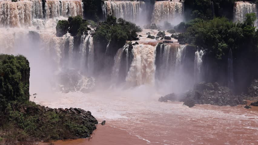 Aerial view of Iguazu Falls with powerful cascades of brown water and mist over rocky cliffs and lush vegetation in Foz do Iguacu, State of Parana, Brazil.
