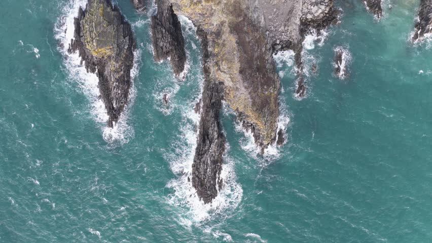 Aerial view of the rugged Welsh Coast with jagged dark rocks and white sea foam crashing against the turquoise water in St Davids, Wales, United Kingdom.