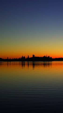 Vertical video capturing a colorful sunset over a large lake in a Canadian national park. Golden and orange hues reflect across the calm water, creating a serene wilderness landscape.