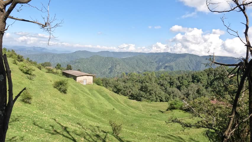 Beautiful panoramic view of green mountain meadows, dense forests, and distant Himalayan hills under a bright blue sky with dramatic clouds. A peaceful countryside landscape showcasing the natural beauty of the Indian Himalayas.