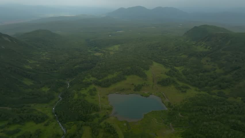 Drone footage of the Vachkazhets mountain range with lush green hills, forested valleys, dirt road and small lake under cloudy sky in Kamchatka Krai, Russia