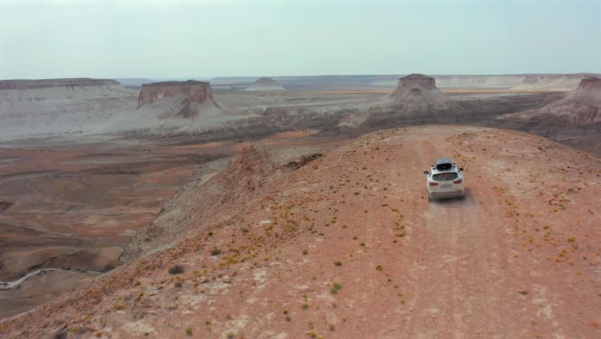 Aerial shot of SUV driving on elevated plateau in Bozzhyra, Mangistau Kazakhstan, overlooking vast desert canyons and ancient seabed formations, highlighting scale, isolation and remote exploration
