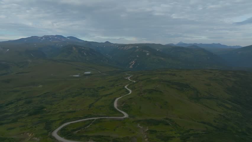 Wide aerial footage of a winding dirt road crossing a vast green valley with mountains and low clouds at dusk in Kamchatka Krai, Russian Federation