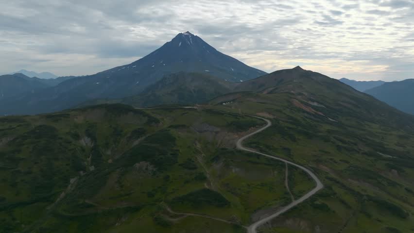 Drone footage of Vilyuchinsky Volcano rising above green ridges and a winding gravel road under a cloudy sky, Kamchatka Krai, Russia