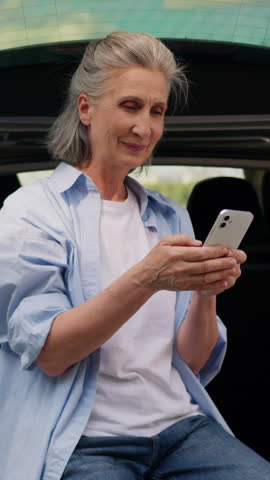 Positive laughing Caucasian old retired woman reading funny news in mobile phone sitting in automobile trunk senior lady aged female with smartphone cellphone gadget device laugh outdoors car travel